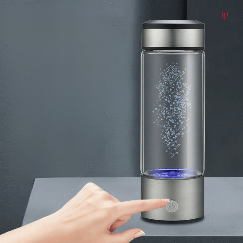 Glass water bottle with silver base and black lid on a dark surface, with a hand touching the base.